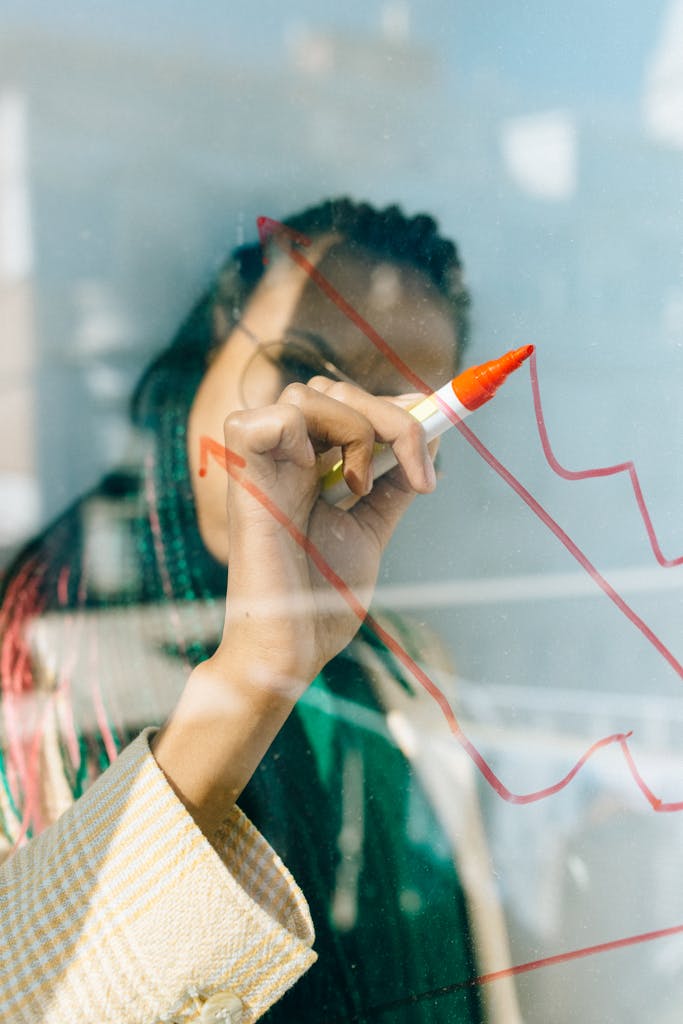 Woman in a Beige Coat Writing on a Glass Panel Using a Whiteboard Marker doing Tax Compliance and Planning