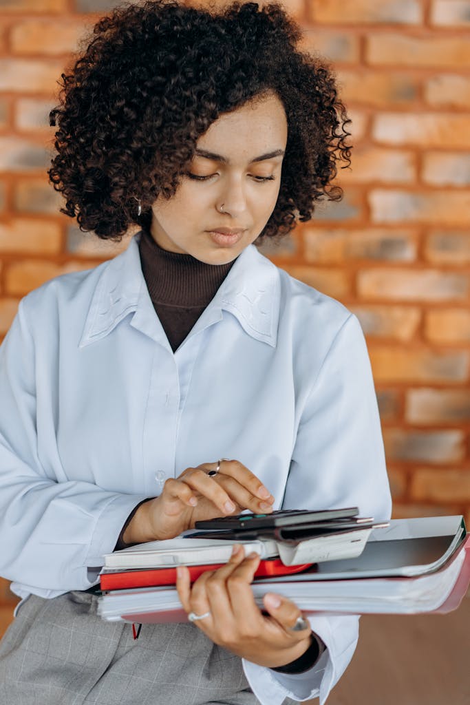 Young Girl in White Long Sleeve Shirt Holding Books and Using a Calculator doing Tax Compliance and Planning